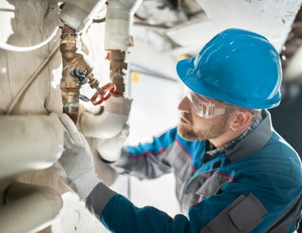 photo of a plumber in a blue hard hat works on pipes in a basement